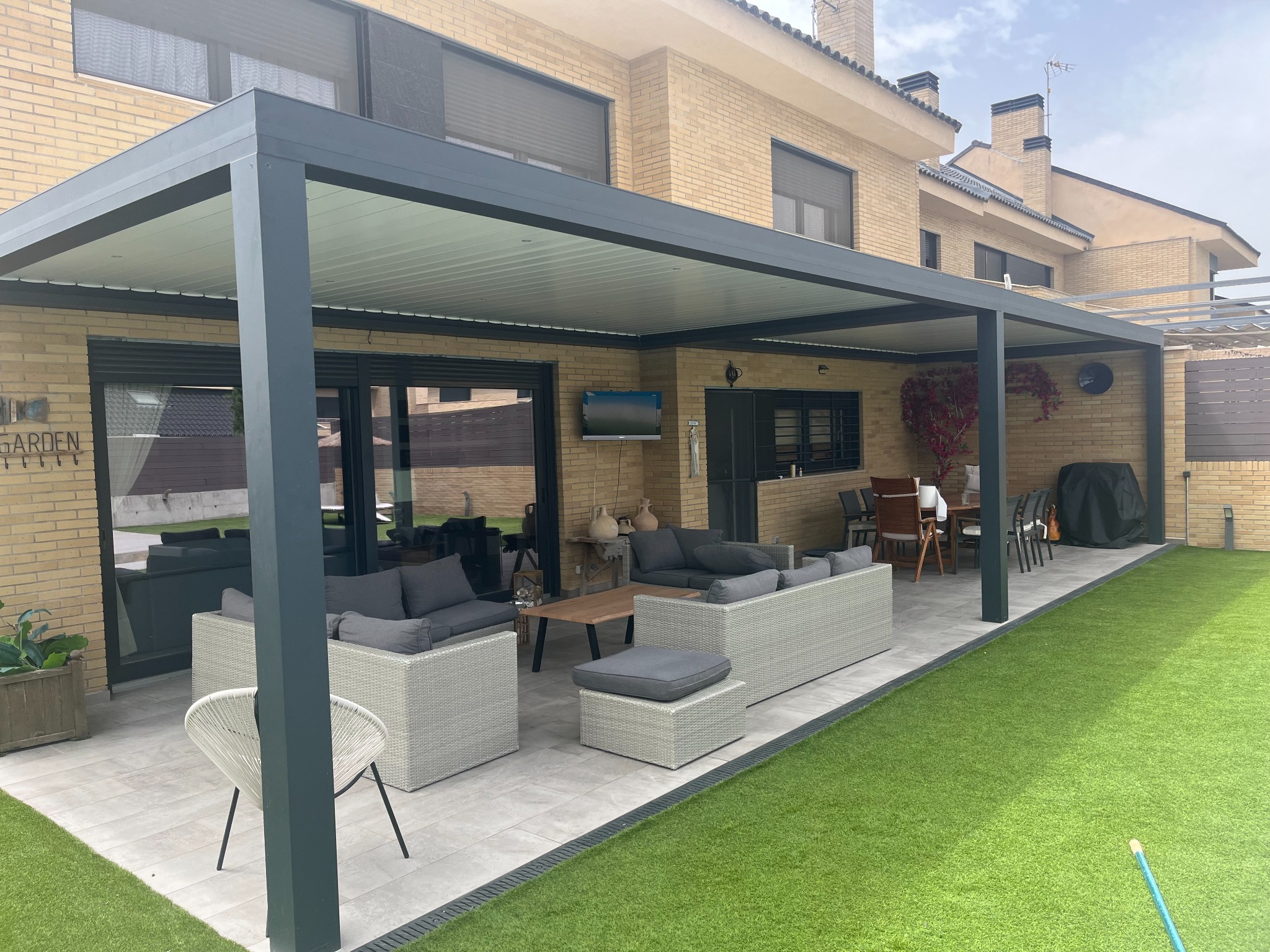 Pérgola bioclimática adosada a una vivienda de ladrillo, con estructura gris y lamas blancas