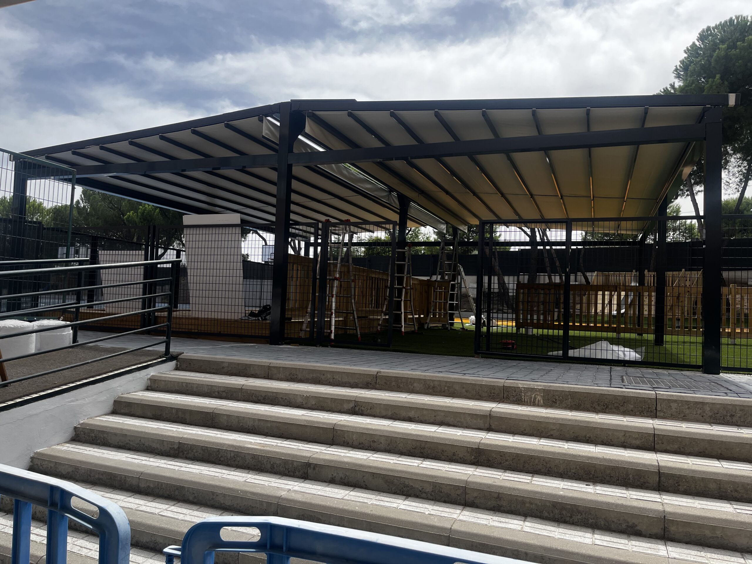 Vista de la pérgola de lona del patio escolar desde el acceso con rampa y escaleras