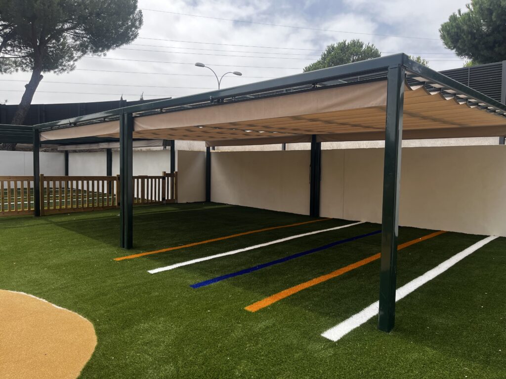 Pérgola de lona beige y estructura verde sobre una zona de césped del patio escolar